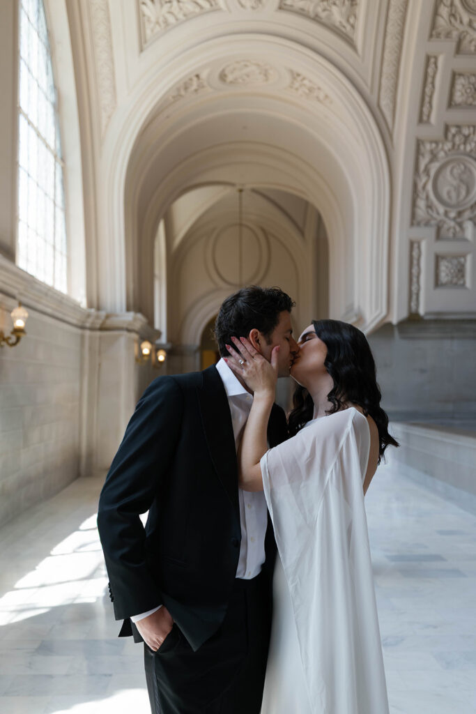 Engaged couple sharing a passionate kiss in the marble hallway of San Francisco City Hall, tall arched windows flooding the corridor with natural light behind them