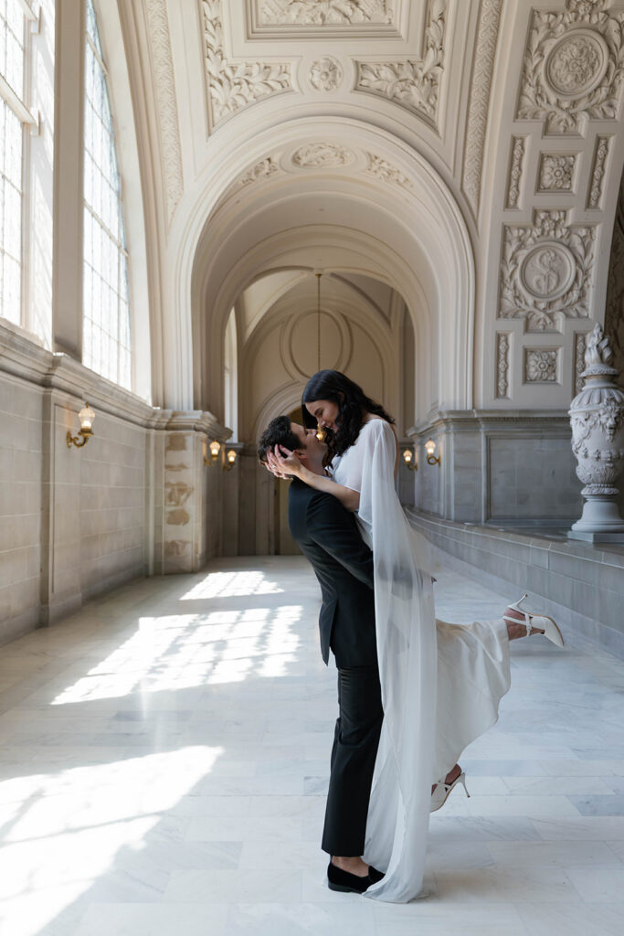 Engaged couple sharing a dramatic lifted kiss in the grand marble hallway of San Francisco City Hall, bride's white cape gown flowing as her heel pops, sunlight streaming across the floor beneath ornate arched ceilings